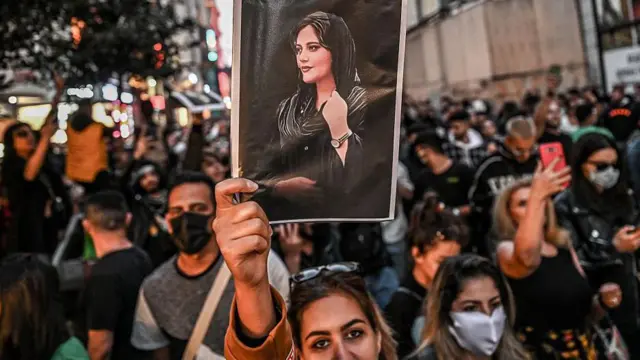 Una mujer mira a la cámara y alza un retrato de la joven Mahsa Amini, en la que lleva velo. 