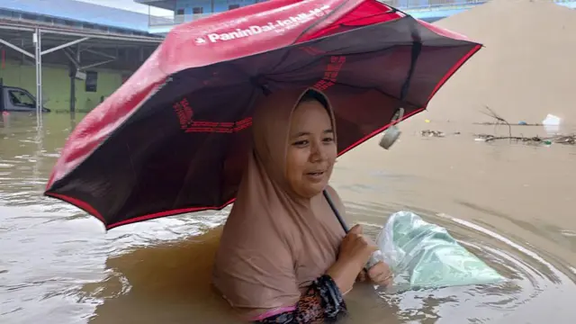 একজন নারী বুক সমান বন্যার পানিতে ছাতা মাথায় দাঁড়িয়ে আছেন, তার হাতে একটি পলিথিনে মোড়ানো প্যাকেট