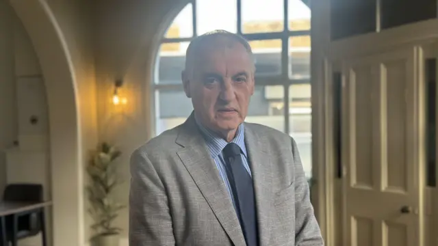 Un hombre con cabello gris y un traje gris claro con una camisa a rayas azul claro y corbata azul marino se encuentra en una habitación color crema con ventanas detrás de él.