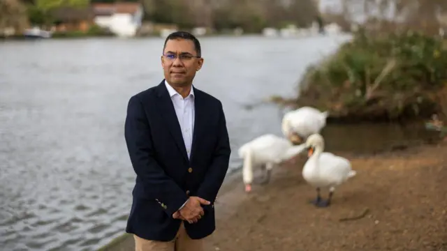 একটি লেকের পাড়ে দাঁড়িয়ে আছেন কালো কোট, সাদা শার্ট ও বাদামি প্যান্ট পরা তারেক রহমান। পেছনে ঘোলাটে ব্যাকগ্রাউন্ডে তিনটি রাজহাঁস দেখা যাচ্ছে 