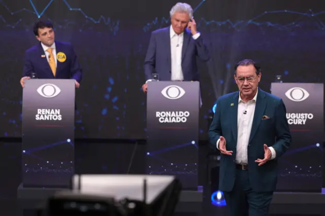 Augusto Cury em debate; ao fundo, desfocados, estão Renan Santos e Ronaldo Caiado