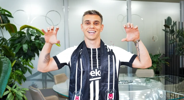 Siyah beyaz Beşiktaş atkılı bir futbolcu gülere poz veriyor. Ellerini kartal tutuşu şeklinde iki yana açmış