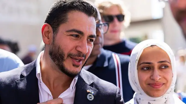 Sakalsız kravatsız siyasetçi ve yanında beyaz başörtülü eşi var. Birilerinin sorusunu adam yanıtlıyor