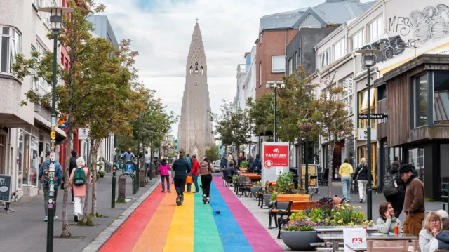 Deptak pomalowany w kolory tęczy, prowadzący do charakterystycznej budowli Hallgrímskirkja (luterański Kościół Hallgrímura). Po obu stronach deptaku rosną rzędami drzewa; chodnikami spacerują małe grupy ludzi. W lewym dolnym rogu widoczne kawiarniane stoliki. W tle strzelista wieża kościelna z szarego kamienia.