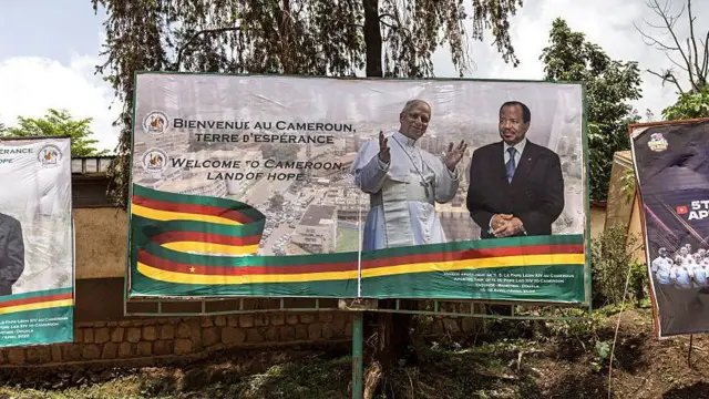 Au centre, un panneau d'affichage arbore le slogan "Bienvenue au Cameroun, terre d'espoir" à gauche, et un ruban à rayures horizontales vertes, rouges et jaunes traverse la moitié inférieure du panneau. Au centre droit, le pape lève les bras, et à côté de lui se trouve le président.