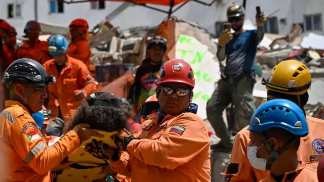 Profissionais de proteção civil, com uniformes laranja e capacetes. Dois deles transportam uma pessoa em uma maca, no local onde ficava um edifício que desabou