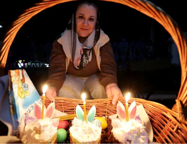 Devojka sa maramom preko kose drži drvenu korpu u kojoj je nekoliko kolača, tri sveće i ofarbana jaja