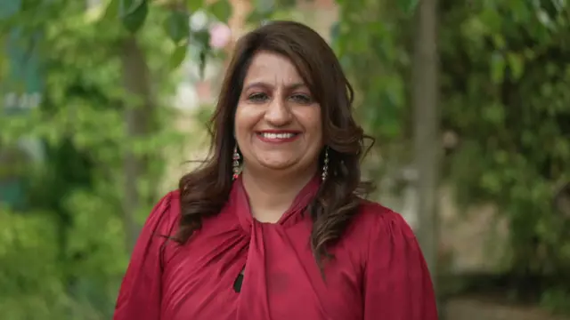 身穿紅色上衣的尼希·吉爾（Nishi Gill）。她戴著彩色耳環，有著棕色捲髮。