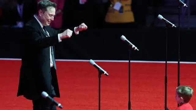 Elon Musk con su hijo, X, en el escenario del mitin de la victoria de Trump en Washington, DC. Musk sonríe y levanta las manos. La cara de X no es visible. 