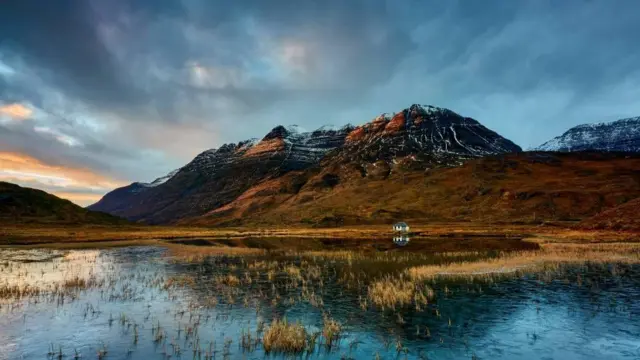 Una pequeña casa blanca reflejada en un pantano helado, con escarpadas montañas al fondo bajo un espectacular cielo nublado.