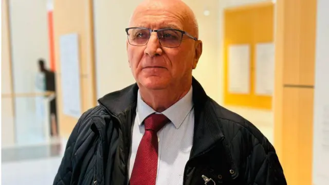 Koloneli Michel Robardey yambaye indorerwamo, ishati y'ubururu bwerurutse na karuvati itukura, n'ikoti ry'umukara rifunguye