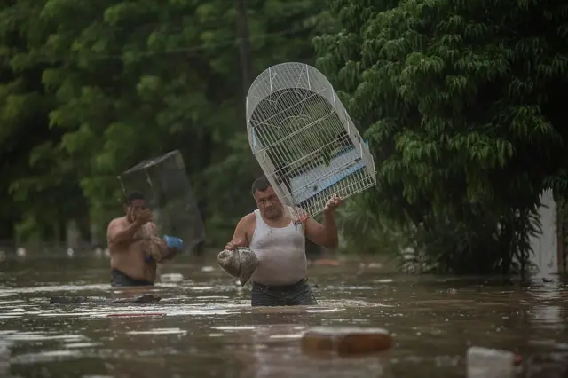 Dos hombres, uno descamisado, con el agua hasta la cintura cargan una jaulas de pájaros y unas bolsas plásticas, huyendo de la inundación