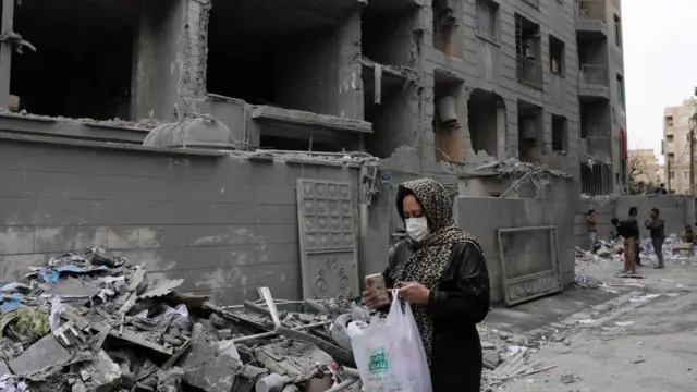 がれきの前を、マスクをした女性がスマートホンを見ながら歩いている。手には白いプラスチックバッグを持っている