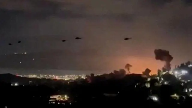 మునుపటి వైమానిక దాడుల నుండి పొగలు స్పష్టంగా కనిపిస్తున్న కారకాస్ మీదుగా ఎగురుతున్న US సైనిక హెలికాప్టర్లు