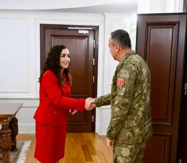 Osmani kırmızı bir döpiyes giyinmiş, kamuflaj giyinen Tümgeneral Özkan Ulutaş ile makamında el sıkışıyor. 