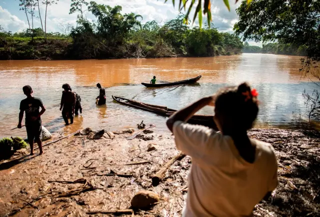 Una mujer de espaldas ve a unas personas en la orilla de un río. En el fonde se ven dos canoas