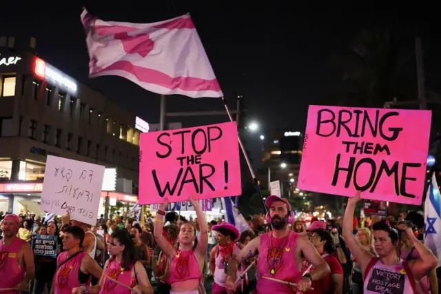 Temmuz 2025'de Tel Aviv'deki protestolarda pem giyimli kadın ve erkekler pembe bayraklar ve Savaşa Bitir, Onları Eve Getir yazılı posterler tutuyorlar. 