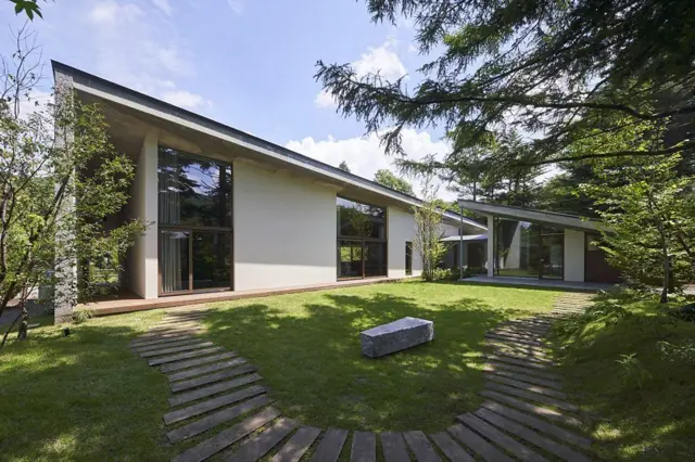 Casa moderna de una planta rodeada de árboles, con grandes ventanales, jardín de césped y senderos de madera curvos.