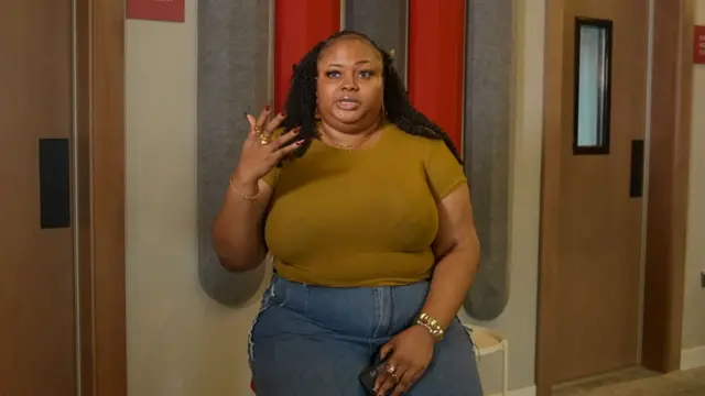 Jennifer wear brown top and blue jeans, camera snap her as she raise her hand dey tok.