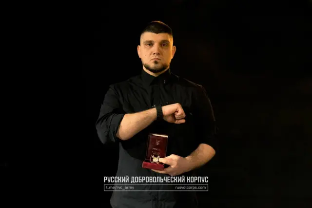 Чоловік у чорному одязі на темному тлі. Він дивиться прямо в камеру з серйозним виразом обличчя. Однією рукою він тримає відкрений футляр із медаллю чи нагородою, а іншою робить жест, ніби показує її або звертає на неї увагу. Освітлення спрямоване з фронту, вирізняючи фігуру на тлі темряви.