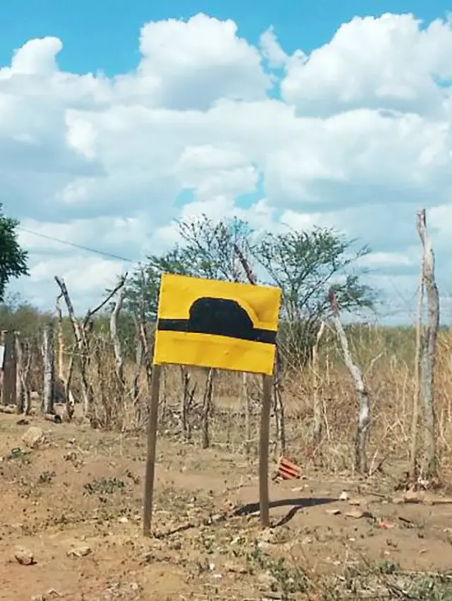 Placa de lombada pintada à mão, fotografada em Francisco Santos, no Piauí. Em volta da placa, se vê uma paisagem de sertão e um céu azul com nuvens