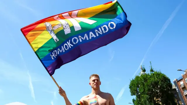 Мужчина с голым торсом держит радужный флаг с надписью Homokomando