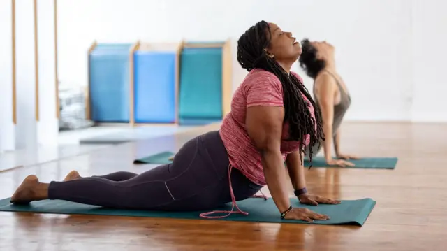 Une femme prend la pose du cobra dans un studio, tandis qu'une autre femme prend la même pose derrière elle. Ses mains sont posées sur un tapis vert et elle regarde le plafond en fermant les yeux.