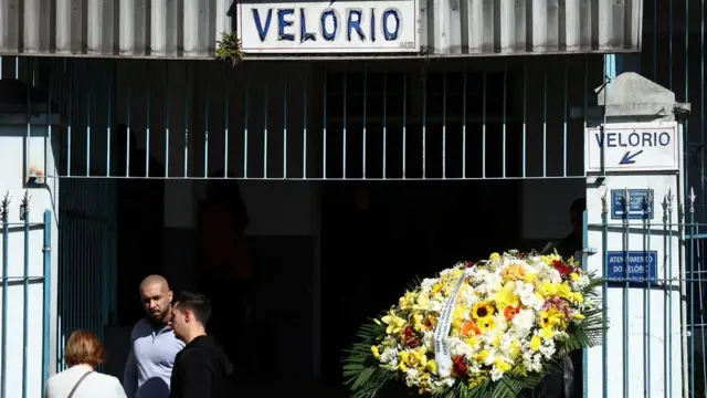 A imagem mostra um local com uma placa escrito "velório", com uma coroa de flores do lado direito e três pessoas conversando do lado esquerdo. 