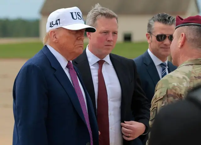 Трамп у костюмі та білій кепці з написом “USA” стоїть на відкритому майданчику поруч із Дрісколлом (у чорному костюмі з червоною краваткою). Перед ними військовий у камуфляжній формі та бордовому береті. На задньому плані видно ще одну людину в костюмі та будівлю з двосхилим дахом.