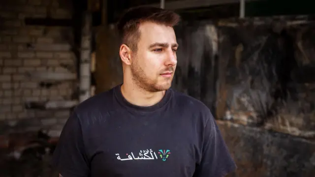 Dmytro Olifirenko lleva una camiseta oscura con un logotipo garabateado en blanco y un tatuaje parcialmente visible en un brazo.