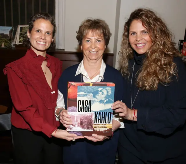 A la izquierda, está Frida Hentschel, quien lleva una blusa roja. En el centro, está Mara Romeo Kahlo, quien viste una blusa blanca y un chaleco oscuro. A la derecha, está Mara de Anda, quien está vestida de negro. Las tres sostienen un libro cuya portada dice "Casa Kahlo" y tiene una foto de Frida Kahlo acostada. Las tres sonríen.