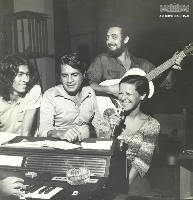 Elis Regina sorrindo ao lado de homens, a maioria sentados; é possível ver alguém ao piano e homem em pé com violão
