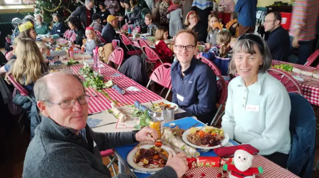 Una fotografía que muestra a personas sentadas alrededor de varias mesas comiendo una cena de Navidad.