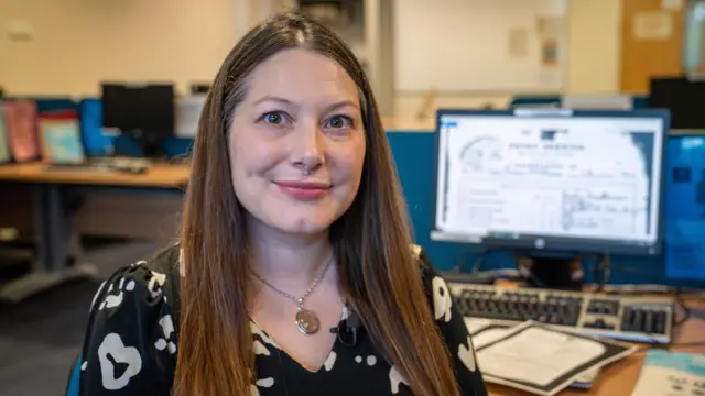 Nicola Nash, sentada em um escritório repleto de livros e documentos. Ela veste uma blusa branca e preta e tem cabelo castanho longo. Está sentada em frente a um computador de mesa, com um documento histórico na tela.
