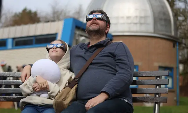 پدر و دختر با عروسکی سفید در بغل روی نیمکت پارک در حال تماشای خورشید‌گرفتگی