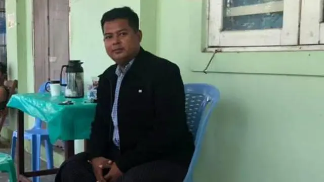 NUG ရဲ့ မန္တလေးတိုင်း ပကဖ တာဝန်ခံ ကိုဝဏ္ဏအောင် (ပုံဟောင်း)၊ အနွေးထည် အနက်ရောင် ဝတ်ထားတဲ့ ကိုဝဏ္ဏအောင်ကို အဆောက်အဦ တစ်ခုထဲ ထိုင်နေတာ မြင်တွေ့ရ၊ ပလက်စတစ်အပြာရောင်ခုံပေါ်မှာ ထိုင်နေပြီး သူက ကင်မရာကိုကြည့်နေ၊ အနောက်မှာ အစိမ်းရောင်နံရံကို မြင်ရပြီး ပိတ်ထားတဲ့ပြတင်းပေါက် တစ်စိတ်တစ်ပိုင်းကိုလည်း တွေ့ရ၊ ဓာတ်ပုံ ဘယ်ဘက်ခြမ်းမှာ အစိမ်းရောင်ပလက်စတစ်အခင်းခင်းထားတဲ့ စားပွဲခုံရှိနေပြီး အဲဒီပေါ်မှာ ဓာတ်ဘူးနဲ့ ပစ္စည်းအချို့ကိုမြင်တွေ့ရ 