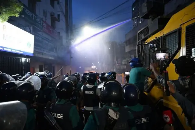 পুলিশের বেশ কিছু সদস্য রাস্তায় দাঁড়িয়ে আছে এবং পুলিশের জলকামান থেকে জাতীয় পার্টির অফিসের দিকে নীল রংয়ের পানি ছোঁড়া হচ্ছে 