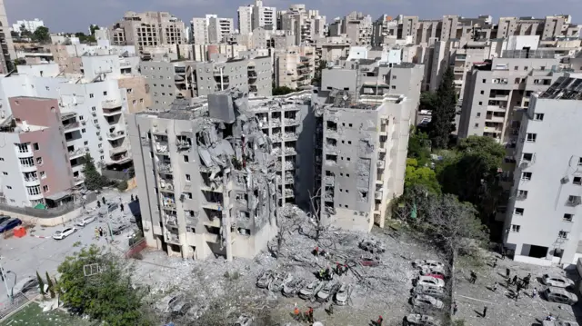 Zdjęcie przedstawiające zniszczony blok mieszkalny i spalone samochody zaparkowane przed nim.