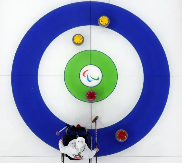 Майданчик для керлінгу з колами синього, зеленого й білого кольорів та паралімпійським символом у центрі – вид згори. Жовті й червоні камені розташовані навколо кола, а внизу кадру спортсмен у кріслі колісному готується зробити кидок.