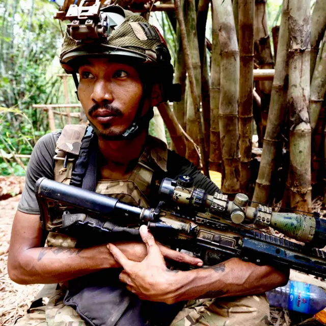 A soldier in a camouflage helmet holds a gun in a jungle setting, a tree trunk behind him. He has a watchful expression and his eyes are fixed beyond the camera.