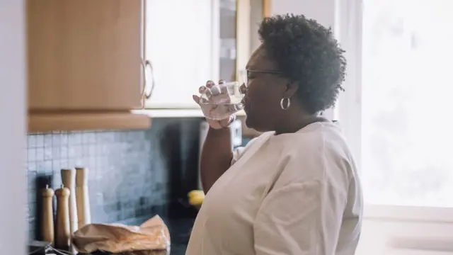 یوه ښځه چې سپین کمیس یې په تن دی پخلنځي کې ولاړه ده او اوبه څښي. شاته یې د اشپزخانې المارۍ او یو څه لوخي ښکاري.