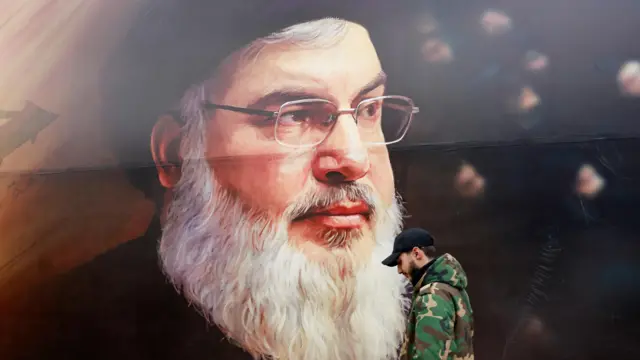 رسم بورتريه لحسن نصر الله بلباسه الأسود المعروف، وأمامه شاب يرتدي لباساً يشبه الزي العسكري وطاقية رياضية سوداء على رأسه