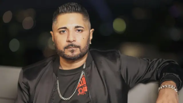 Jash Negandhi sentado em um sofá, com camiseta e jaqueta pretas