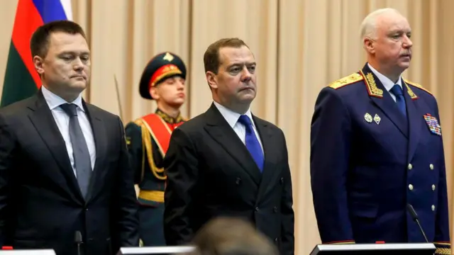 Краснов, Медведев и Бастрыкин стоят на каком-то совещании на фоне военнослужащего в парадной форме и флага России