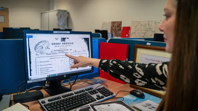 Nicola Nash, sentada em um escritório, rodeada de livros e documentos. Ela usa uma blusa branca e preta e tem cabelo castanho longo. Ela aponta para um computador de mesa com um documento histórico na tela, que é o histórico de serviço do soldado Gordon McPherson.