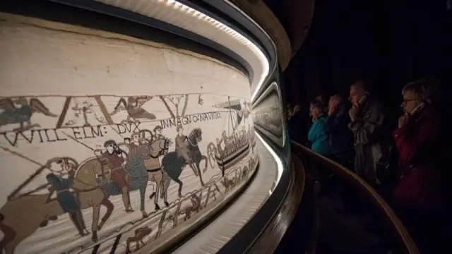 横に細長い布に、馬やボートに乗った兵士などが刺繍されている。布はガラスケースに入れられており、美術館の訪問客がそれを眺めている