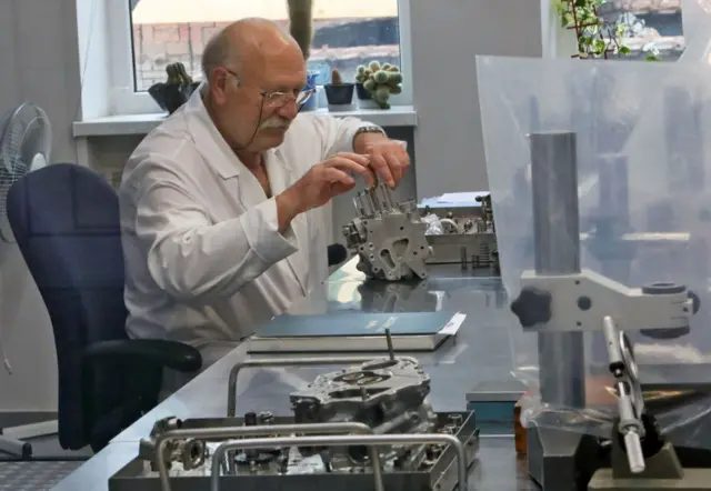 Технічна лабораторія або майстерня. На передньому плані – металевий стіл із деталями складних механізмів, інструментами та вимірювальним обладнанням. У центрі людина в білому халаті працює з великою металевою деталлю, схожою на частину двигуна. На столі також лежить відкрита книга та кілька інших компонентів. У фоні видно вікно з горщиками з кактусами.