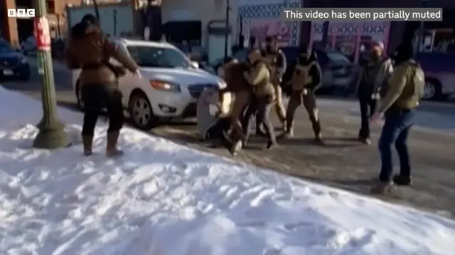 На зимовій вулиці, де лежить сніг, кілька озброєних людей у тактичному одязі силоміць затримують когось біля білого автомобіля. Одна людина стоїть осторонь, спостерігаючи. Сцена виглядає хаотичною й напруженою.