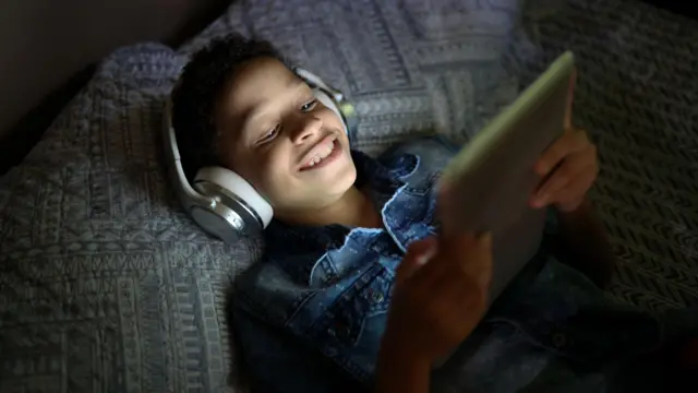 Criança sorrindo enquanto assiste algo na cama, com table e foneouvido