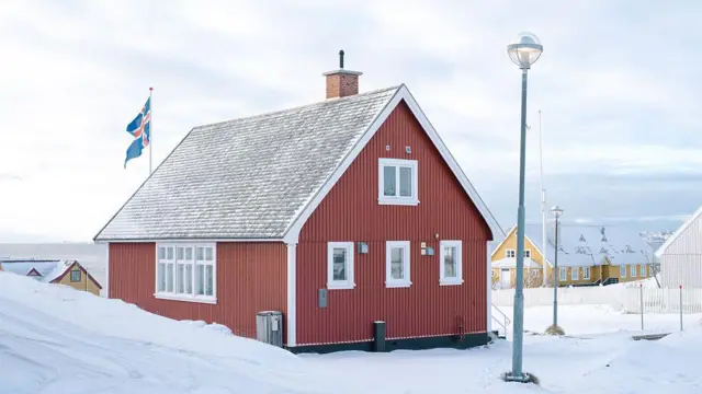 Одноэтажный домик цвета терракоты среди снега. На крыше - флаг Исландии